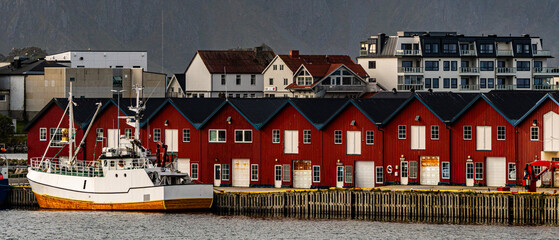 Traditional red wooden fishing huts and white boat in a scenic nordic harbor beneath a steep dark mountain wall with copy space © Quality by Simon