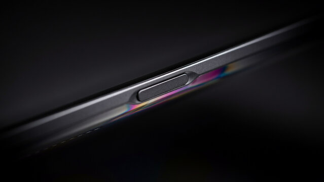 A macro view of a metallic smartphone side frame featuring a power button and a vibrant rainbow light refraction on the dark gray surface
