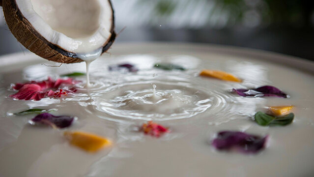 Coconut milk being poured from a cracked coconut shell into a bowl filled with white liquid, colorful flower petals, and creating circular ripples