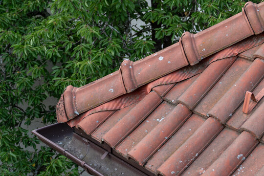 Red ridge cap on the corner of a roof with red tiles, a gutter, and a tree in the background.