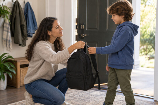 Mother helps a boy prepare his backpack at the home entrance. They share a calm school morning routine with kind guidance and organization. Family care concept for ADHD awareness.