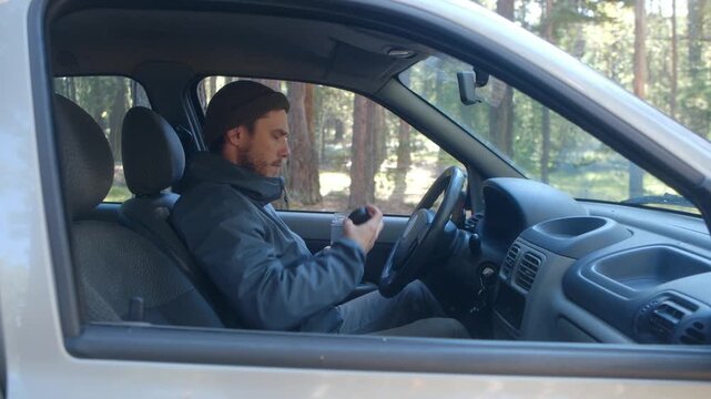 Young bearded man in a beanie and jacket sitting inside his car in a pine forest, taking a break to open a plastic bottle and drink fresh water, quenching his thirst during a road trip