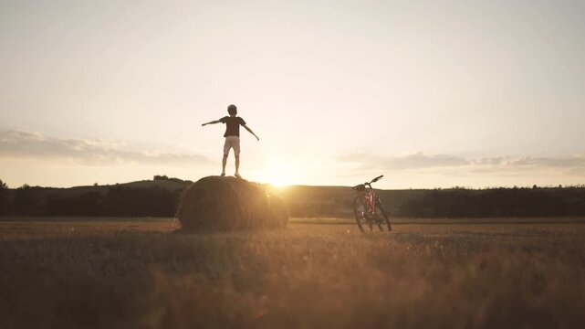 Child boy admiring beautiful sunrise or sunset in countryside, stand on haystack . Carefree schoolboy enjoying merry vacation or holidays in nature, picturesque rural scenery, farming and agriculture