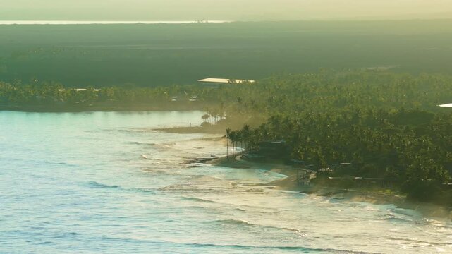 Aerial drone view of Big Island Hawaii coastline at golden sunrise with turquoise waves breaking on reef dense palm groves misty backlit light and small beachfront houses along tropical shore.