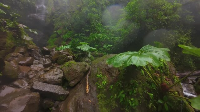 FPV drone flight through misty rainforest canyon in Costa Rica revealing tall waterfall plunging over cliff into turquoise river surrounded by lush green tropical jungle and mossy rocks.
