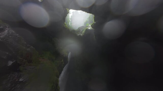 FPV drone flight through narrow misty canyon in Costa Rica rainforest with moss covered rock walls waterfall mist and bright light breaking above lush tropical green vegetation and wet stone.