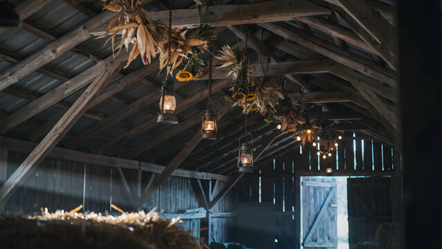 Rustic barn interior with hanging dried corn and sunflowers, vintage lanterns suspended from wooden beams, and a pile of golden hay in front