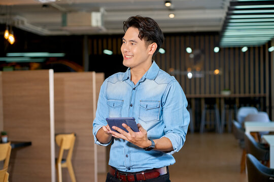 Smiling young Asian man in denim shirt holding digital tablet in modern coworking space. Friendly tech-savvy entrepreneur or freelancer standing in office.