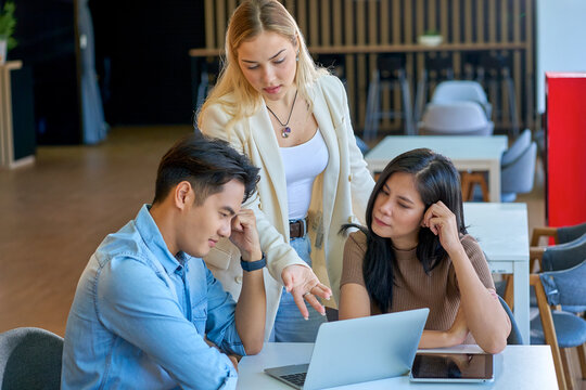 Diverse creative business team discussing strategy on laptop in modern coworking office. Female leader explaining project ideas to attentive colleagues at desk.