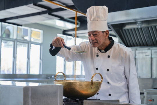 Mature Asian chef tasting soup with ladle in commercial restaurant kitchen. Professional senior chef in toque hat cooking food in brass wok.
