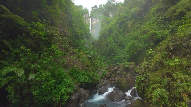 FPV drone flight over rocky river through lush green jungle canyon in Costa Rica with tall misty waterfall falling from cliff into turquoise stream surrounded by ferns and tropical plants.