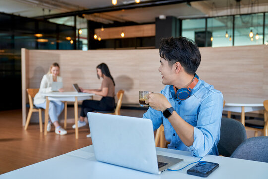 Young Asian man holding coffee cup working on laptop in casual coworking space. Freelancer enjoying work in modern office with colleagues in background.