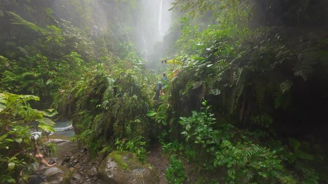 FPV drone flight through lush misty rainforest canyon in Costa Rica revealing father and son hikers on rocky trail beside rushing river and tall waterfall falling into deep green gorge.