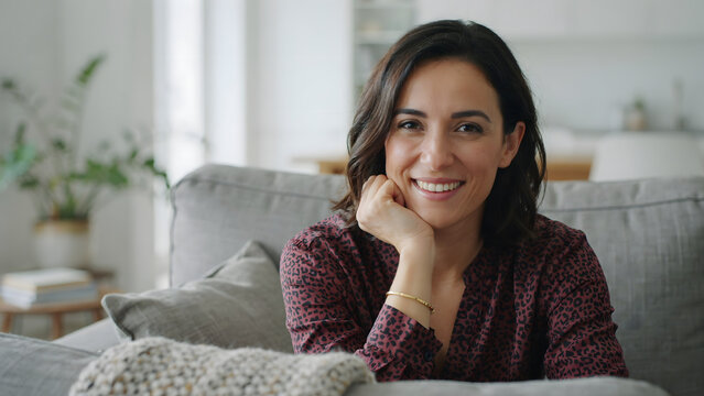 A smiling young woman with dark hair sitting on a comfortable grey sofa in a bright living room, resting her chin on her hand and looking at camera