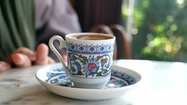 A close up view of a hand lifting a small, ornate turkish coffee cup from its matching saucer on a marble table, showing steam rising from the hot beverage