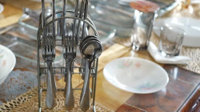 A close up view of a collection of silverware neatly arranged in a metal holder on a wooden dining table, with sunlight casting shadows on the surface