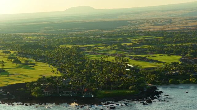 Aerial drone view of a luxury golf course resort on the Big Island of Hawaii with palm groves red roof villas black lava rock shoreline turquoise ocean and warm golden sunset light.