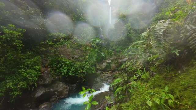 FPV drone glides through misty jungle canyon in Costa Rica where a tall waterfall drops into turquoise river between mossy cliffs and ferns as a father with son explore lush tropical forest.