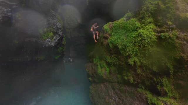 FPV drone view of man descending on rope down mossy cliff into misty canyon with turquoise river in Costa Rica. Rain falls over wet rocks and lush green jungle walls of hidden gorge.