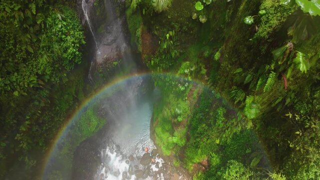 FPV drone flight through misty rainforest canyon in Costa Rica where a group of people enjoying under waterfall stand on rocks beside turquoise river as rainbow glows in the spray.