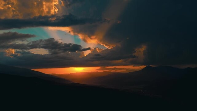 Golden sun peeks through stormy clouds. Silhouetted mountains stretch beneath the sky's drama. Light fractures through heavy, brooding clouds. Dusk's edge glows warmly against the darkened horizon