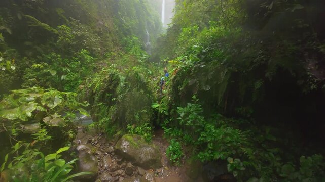 FPV drone flight through misty Costa Rica rainforest gorge where father and son stand on a rock watching a tall waterfall fall past moss covered cliffs and lush green tropical ferns.