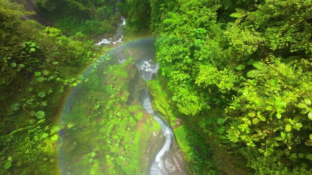 FPV drone flies through lush Costa Rica rainforest gorge with rainbow mist above turquoise river where father and son enjoy bathing under waterfall surrounded by mossy rocks and giant leaves.