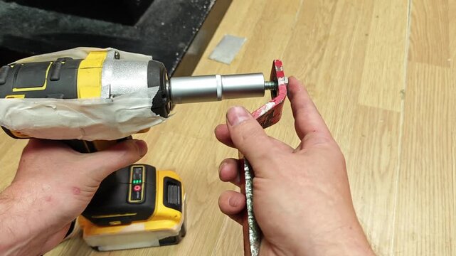 Impact driver and red bracket stay aligned as fastening continues. Hands hold metal part in place while threaded rod extends outward, creating precise tool adjustment task on workbench.