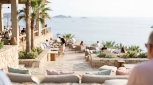 Patrons enjoy a relaxing afternoon at an outdoor seaside restaurant terrace. The scene features soft focus on guests dining near the ocean with palm trees and stone architecture.