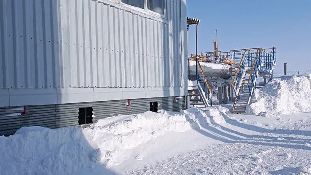 Snowbound industrial facility sits under clear winter sky, with metal stairs, pipes, and heavy snowdrifts outside. Arctic facility exterior feels quiet and isolated in bright cold daylight.