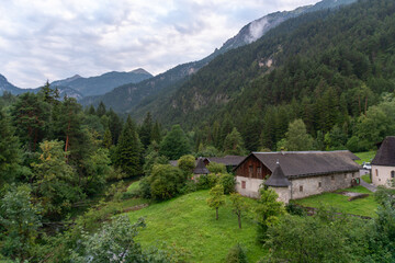 Obraz premium Traditional stone and timber buildings stand in a lush valley near Fernsteinsee in Tyrol, Austria. Forested Alpine slopes, meadow and low mountain clouds create a scenic rural setting for European tra