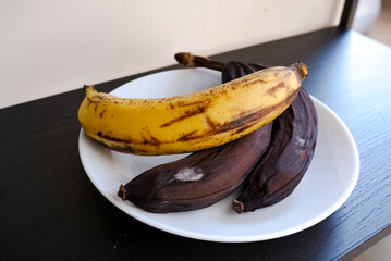 Overripe black bananas on white plate on dark table close up. Concept of food waste, spoiled tropical fruit, ingredients for banana bread baking, rotten fruit, and kitchen still life. © Олена Сосіна