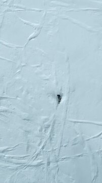 Single snowboarder carving shallow turn on wide wind-sculpted snow slope, clear parallel tracks visible from overhead drone view with light snow spray