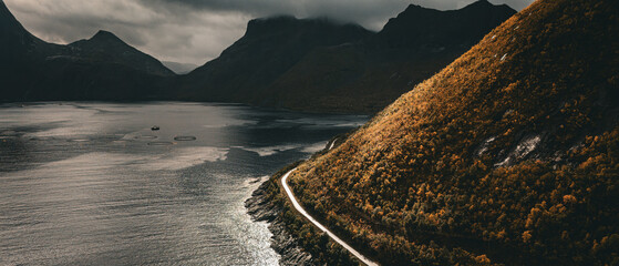 Aerial view of a winding coastal road along a colorful autumn mountain slope and fjord under dramatic sky with copy space © Quality by Simon