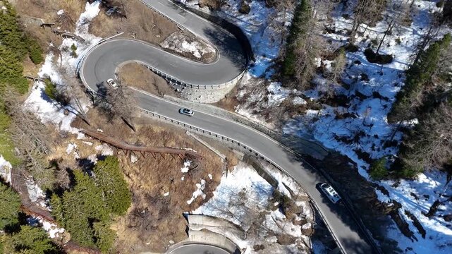 oblique aerial angle winding switchback road stone retaining wall guardrail two cars climb forested slope patchy snow malojaschlange maloja pass graubunden switzerland swiss alps mountain pass