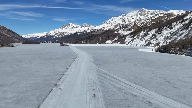 frozen silsersee ice sheet groomed ski track winds toward snowy granite peaks drone flyover engadin valley switzerland larch forest shoreline alpine hamlet clear blue sky winter landscape