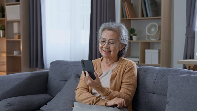 Happy senior Asian woman looks at a smartphone while relaxing on a sofa at home, showing positive aging, independence, digital connection, mobile technology and a comfortable retirement lifestyle.