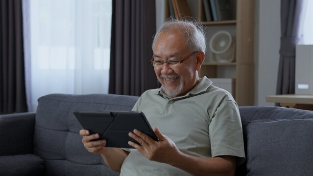 Cheerful elderly Asian man sitting on comfortable couch, holding tablet with both hands and smiling happily while using mobile app in modern living room.