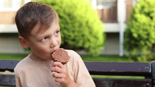 Boy eats chocolate ice cream calmly on hotel bench but shows little enjoyment. Round dessert with chocolate coating and sprinkles seems less appealing to him.