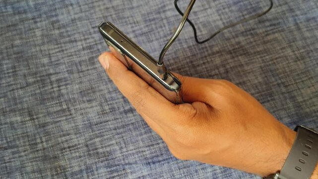 Person hand holding smartphone and plugging or connecting an old traditional wired headphone cable cord male cylindrical pin into round type smartphone female audio signal jack socket or port hole.