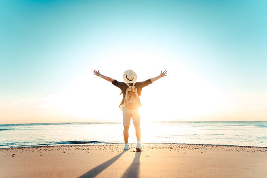 Happy man with backpack standing with arms up at the beach - Delightful tourist enjoying summer vacation by the seaside - Traveling life style and well being concept
