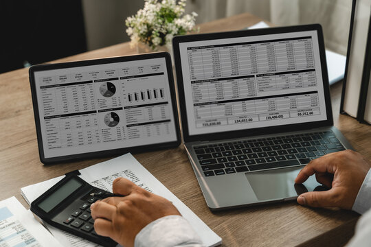Businessman reviewing financial spreadsheets on laptop and tablet while using calculator with printed reports, showing accounting analysis and financial data review.