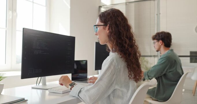 Woman developer coding software on computer monitor. Focused programmer works at desk while colleague uses a workstation in a bright modern office. Tech career and coding project concept.
