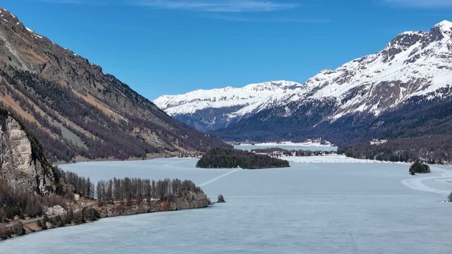 frozen silsersee valley panorama wooded island alpine village distant engadin switzerland snowcapped granite peaks larch forest shoreline drone aerial flight winter view