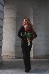 Fototapeta premium Red Paeonia lactiflora flowers held by a woman in a black dress standing within an arched stone corridor