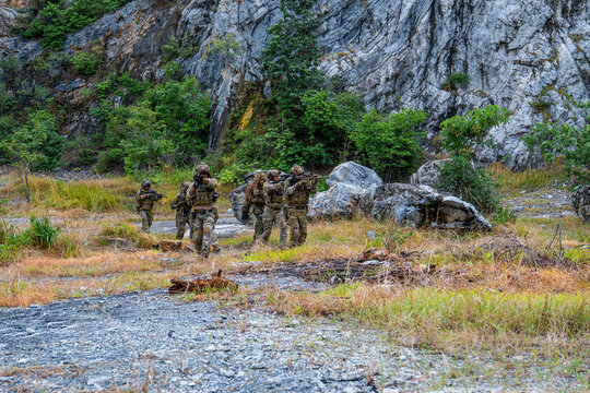 Soldiers move through rough terrain during a training exercise in a forested area near rocky cliffs