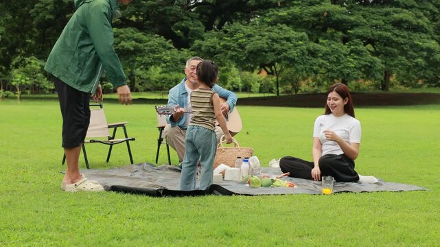 GrandFather playing acoustic guitar for child during picnic. Asian family enjoy outdoor picnic with food and drinks in park sunny day, outdoor activitie with daughter father, mother grandfather