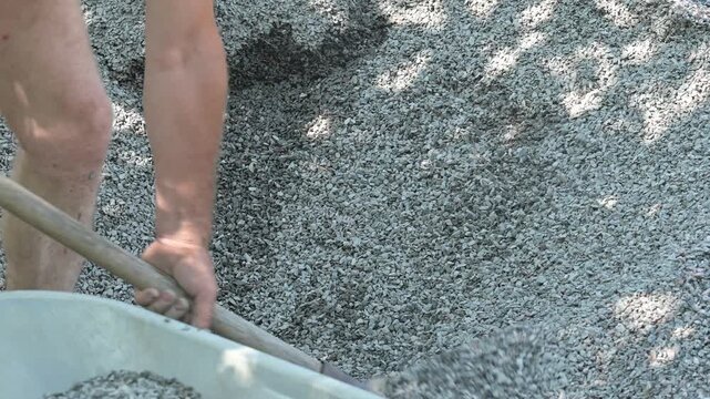 Shovel pouring crushed stone into a wheelbarrow on a construction site. Close-up of a worker's hands with a shovel pouring small gravel into a wheelbarrow, hard physical labor and construction work