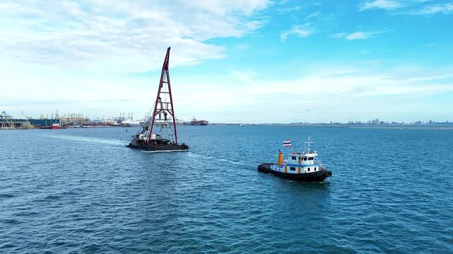 Tugboat towing a heavy crane barge for offshore construction in the blue sea near a commercial port terminal. Marine infrastructure development, maritime logistics, and industrial freight.