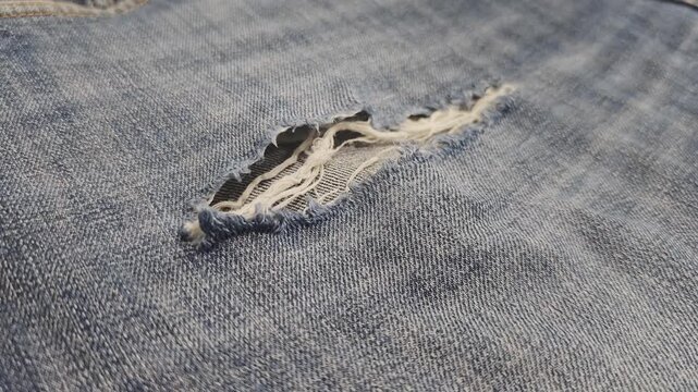 Detailed macro shot of worn blue jeans. The camera slowly pans over a torn hole revealing the texture of the denim and frayed fibers